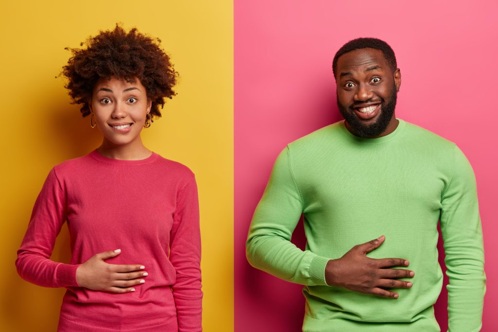 Pleased ethnic young woman and man keep hands on stomach symbolizing easy digestion, feel satiety after eating tasty nutritious dinner, smile positively, happy not to be hungry, pose against yellow and pink wall
